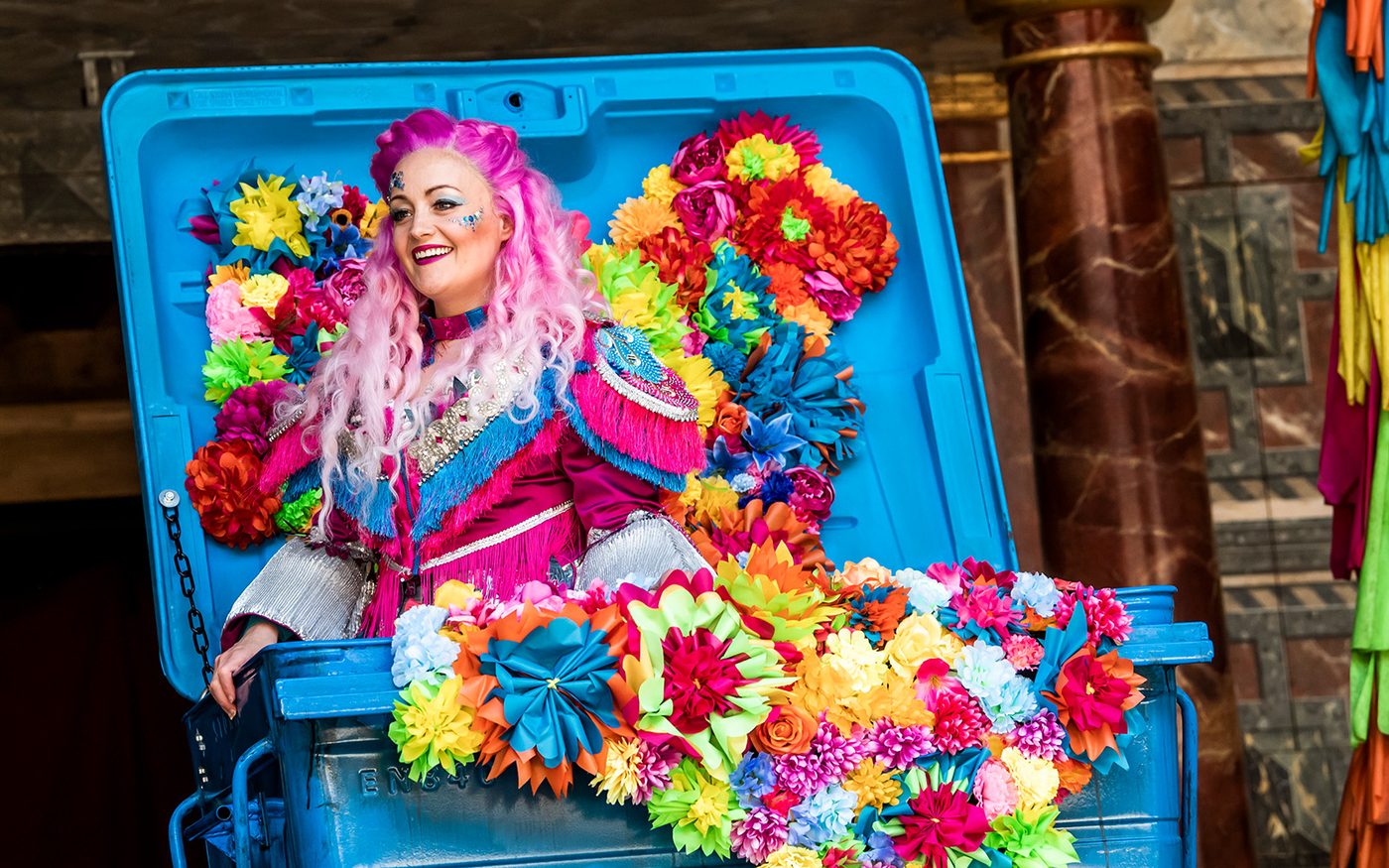 Actor Victoria Elliot portraying the character Titania in bright multi-colored dress resembling flowers emerging from what appears to be an industrial container